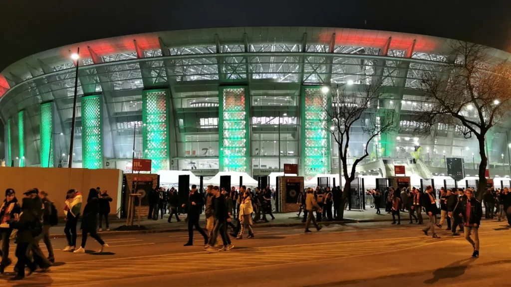 The Puskás Aréna in Budapest at night, its facade lit green and red, with crowds of fans outside.