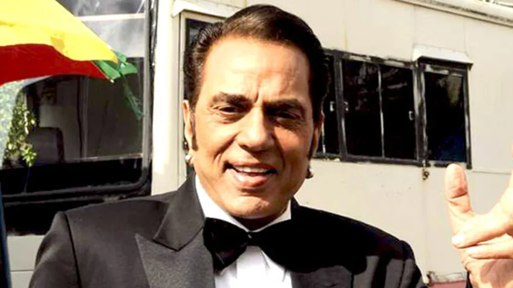 Dharmendra photographed in 2011, in a tuxedo and bow tie, smiling and raising two fingers.