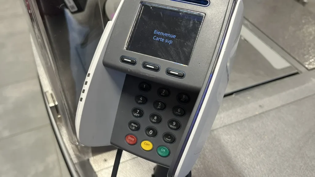 A card payment terminal with a screen and a full numeric keypad, fixed on a stand over a brushed metal checkout counter, with the edge of a clear perspex screen at the left. Illustrative photograph of a stock terminal, not of any business, venue or person named in this article, and it does not show any surcharge being applied.