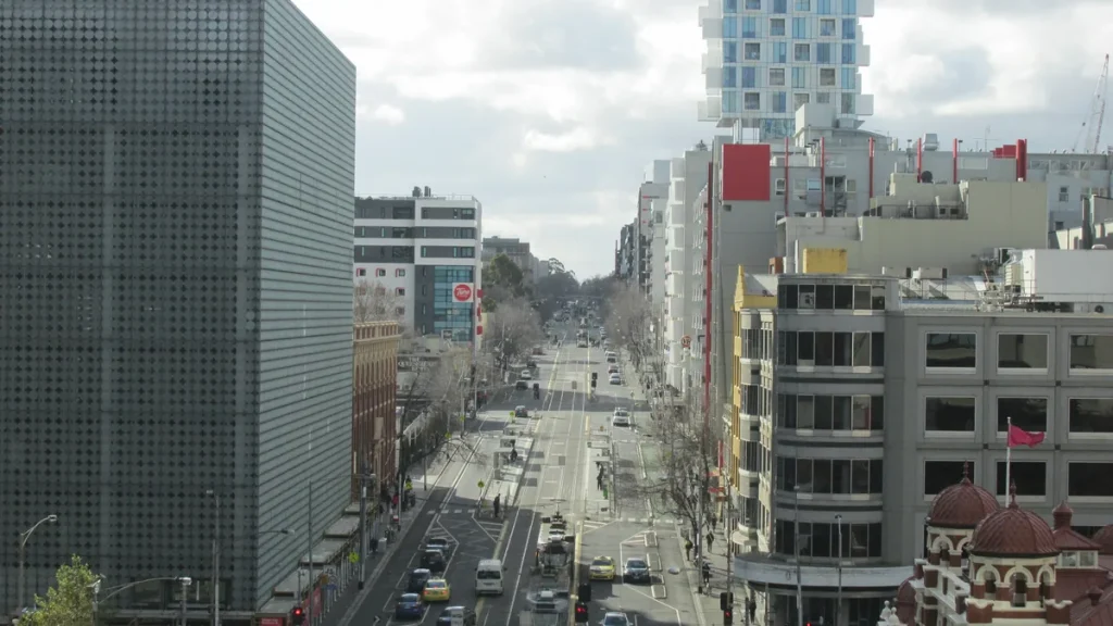An elevated view along a Carlton street in inner Melbourne, with tram tracks, a tram and cars on the road, buildings on both sides and bare winter trees under a cloudy sky. Illustrative photograph of the suburb, not of the address, the scene or any person involved.