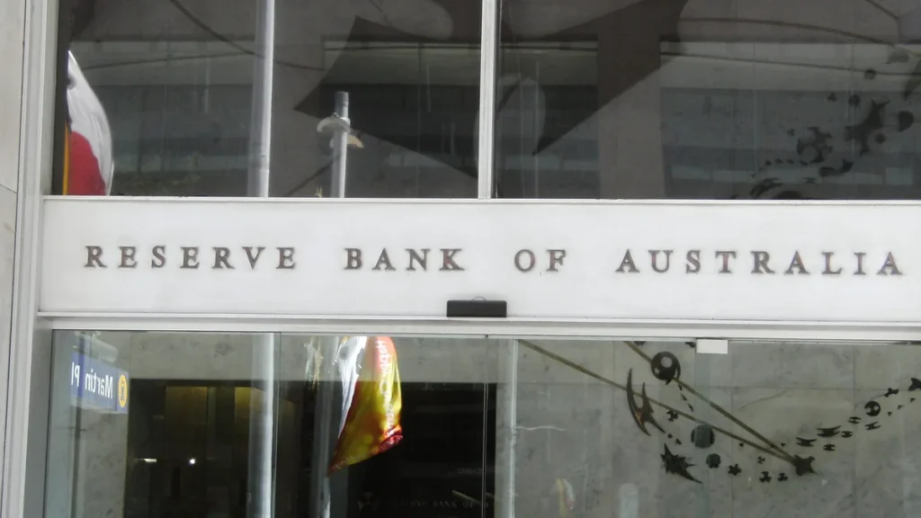 Raised metal lettering reading RESERVE BANK OF AUSTRALIA on the white fascia band above the glass entrance doors of the bank's Martin Place head office in Sydney, with a reversed Martin Pl street sign and other reflections visible in the glass. Photographed in November 2008, not from the September 2026 decision.