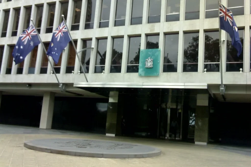 The concrete and glass entrance of 1 Treasury Place in Melbourne, with two Australian flags and a Victorian flag on poles and the Victorian coat of arms on a green panel above the doors.
