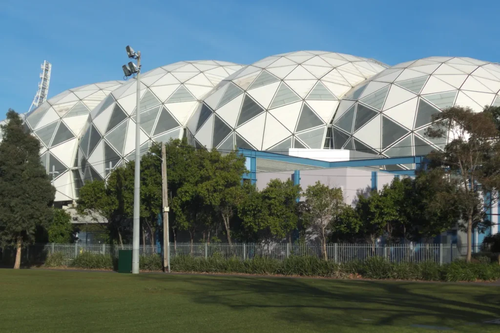 The white geodesic domed roof of AAMI Park in Melbourne rising behind a row of trees and a metal fence, with a light tower at left and a mown lawn in the foreground.