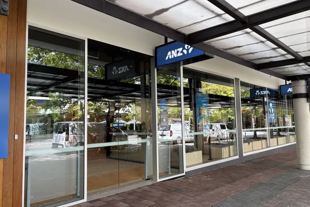 The glass shopfront of an ANZ branch in the Canberra Centre, with the blue ANZ sign hanging above the entrance and a covered footpath of red brick pavers. Photographed in March 2026.