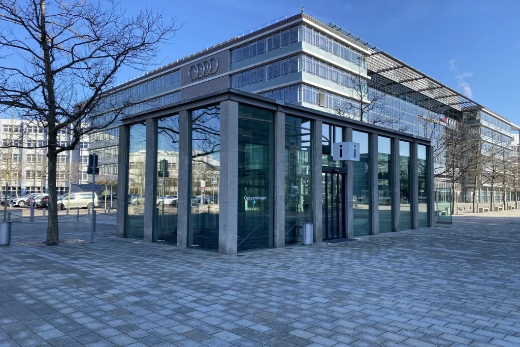 A glass-walled visitor pavilion in front of a multi-storey office block bearing the four Audi rings, under a clear blue sky in Ingolstadt.