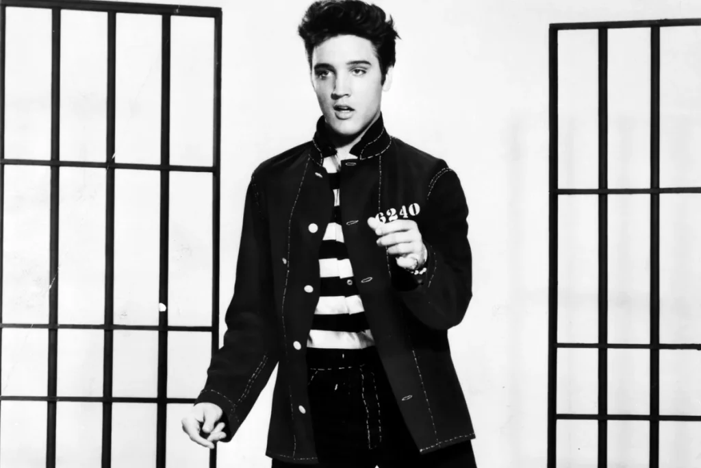 A black and white photograph of a young Elvis Presley in a dark denim jacket over a striped shirt, posed between barred cell doors.