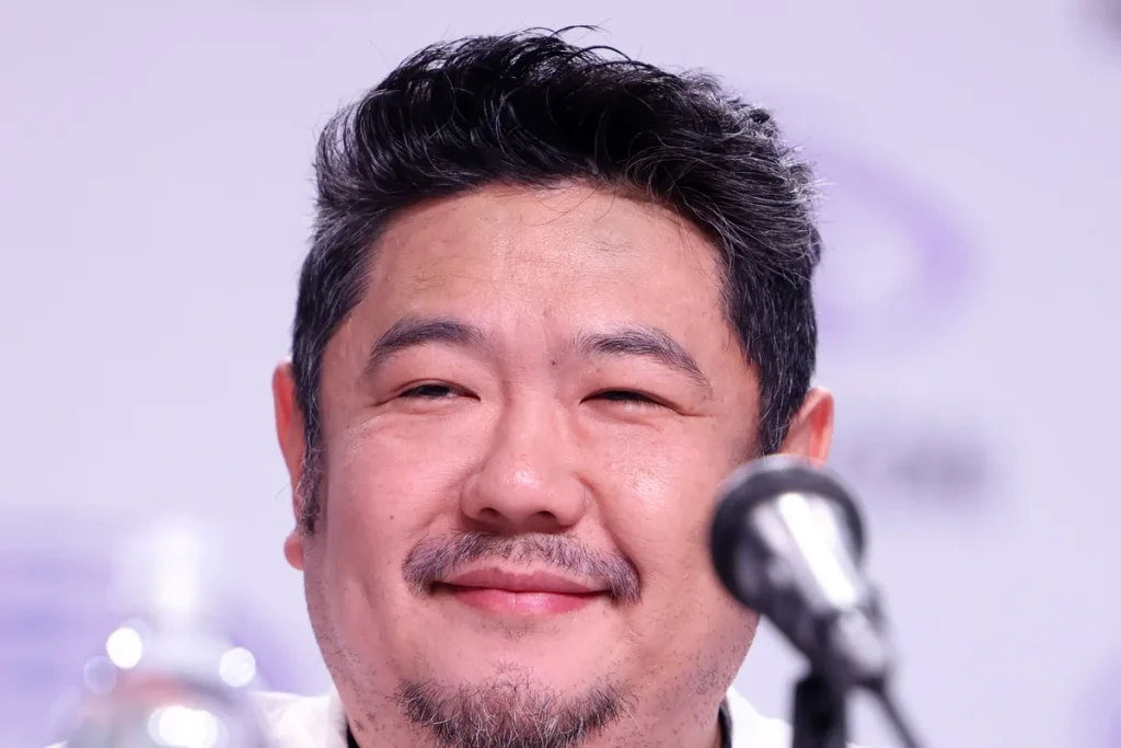 Eric Bauza, smiling, with dark swept-back hair greying at the sides and a goatee, seated behind a microphone against a pale lavender backdrop.