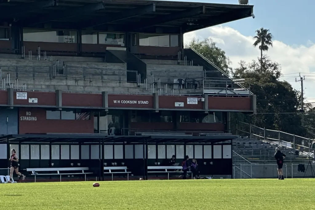 An Essendon player in red and black running across the sunlit oval in front of the old concrete W H Cookson Stand at Windy Hill, with a football resting on the grass.