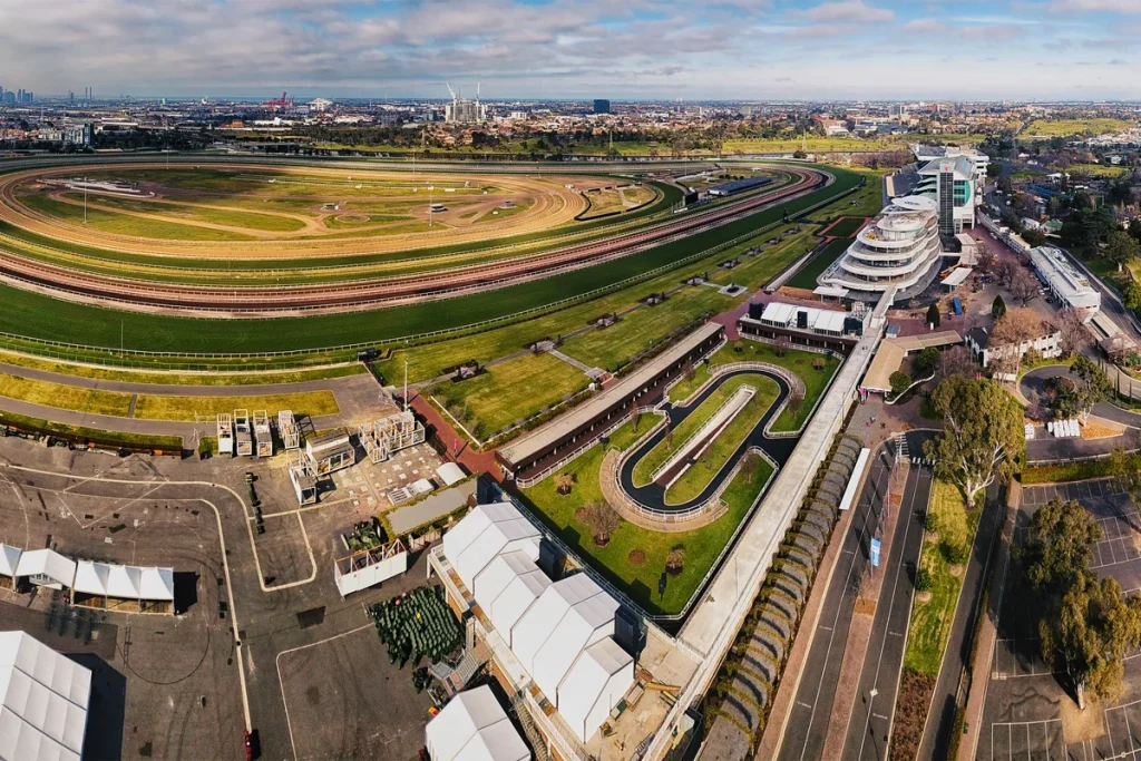 Aerial view of Flemington Racecourse in Melbourne: the green turf track curving around a sand infield, the white tiered grandstand and the horseshoe-shaped mounting yard, with suburbs and low cloud behind.