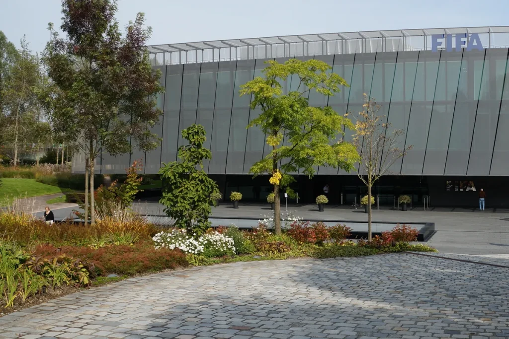 The long grey glass-and-mesh facade of FIFA's headquarters in Zurich with the FIFA name on the roofline, behind landscaped gardens and a cobbled forecourt.