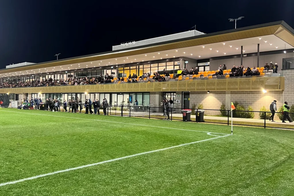 A floodlit football pitch at night in front of a long two-storey grandstand with orange seats and a gold-clad balcony, spectators along the fence and in the stand.