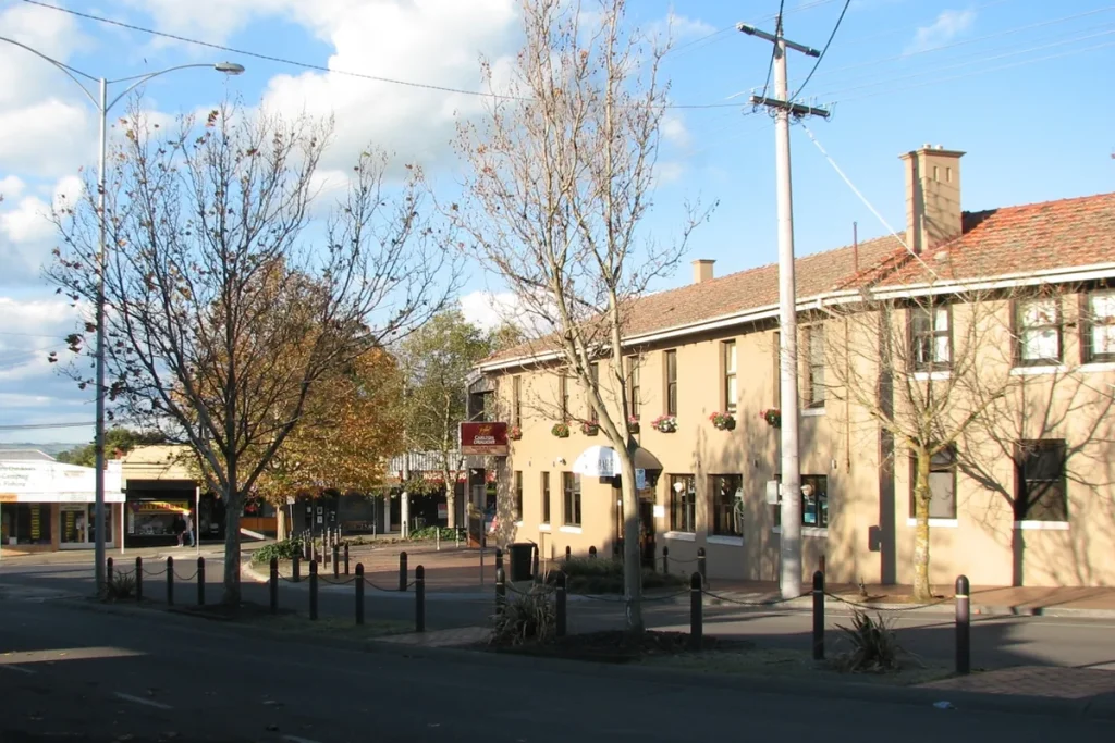 McCartin Street in Leongatha on a sunny day, with a two-storey cream hotel with a red tiled roof and flower boxes on the right, bare and autumn-leaved street trees, and a row of shops along the footpath at the left.