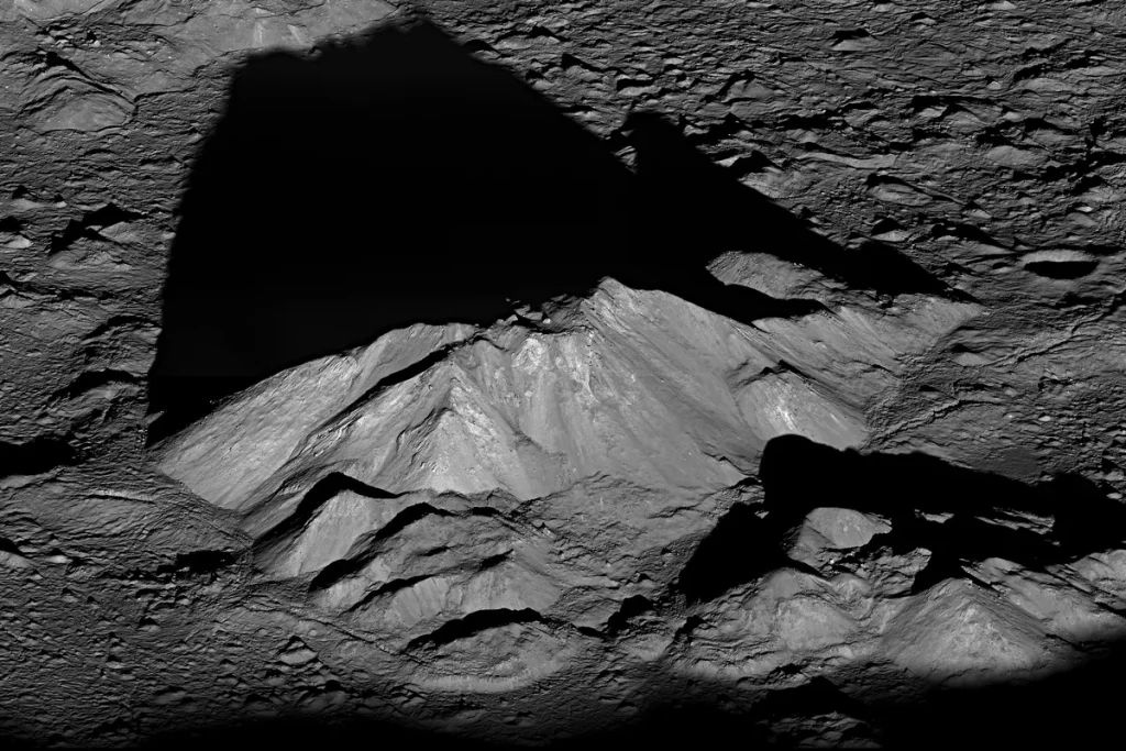 The sunlit central peak of Tycho crater on the Moon seen obliquely from orbit, its long black shadow stretching across the rough, cratered crater floor.