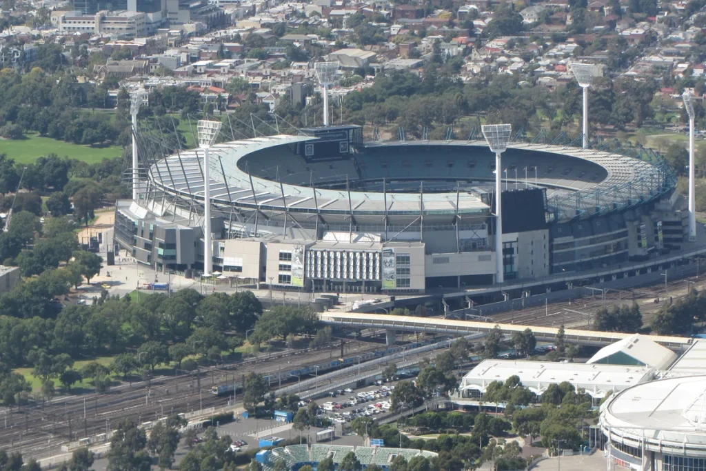 An aerial view of the Melbourne Cricket Ground's oval stands and light towers, with parkland, rail lines and Melbourne's eastern suburbs around it.