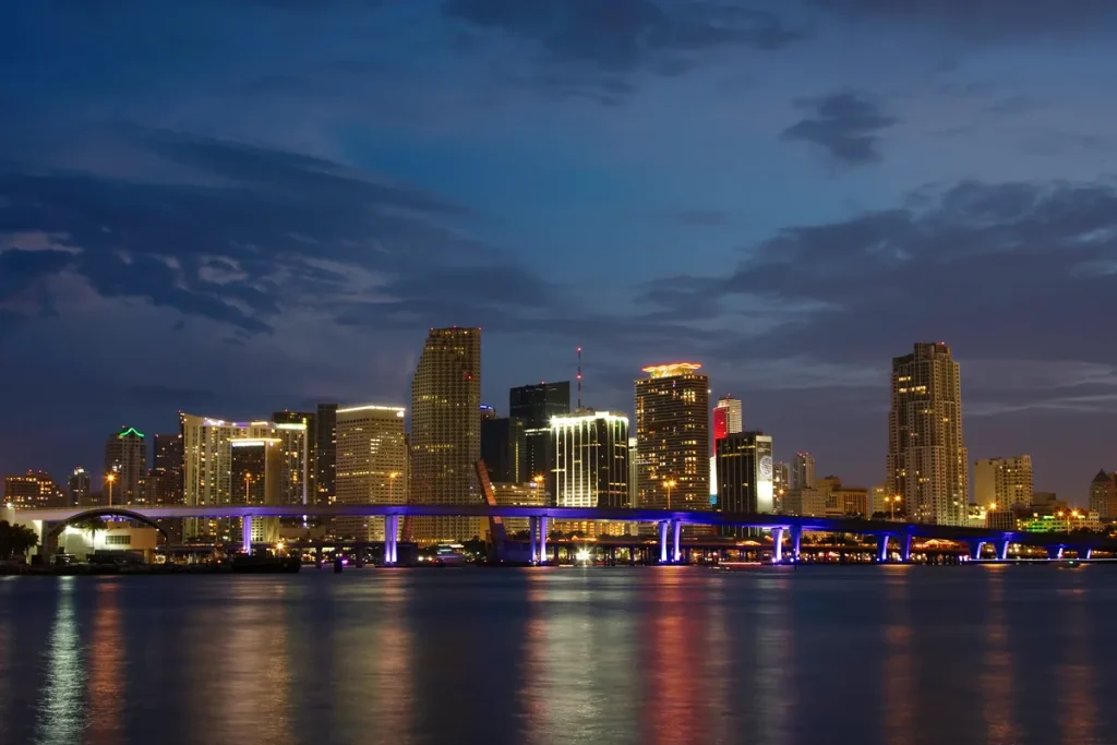 The downtown Miami skyline at dusk across calm water, tower lights reflected in the bay and a causeway lit purple underneath.