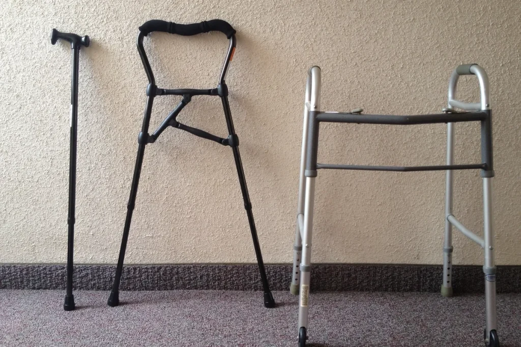 A black walking cane, a black hybrid walking cane and a grey folding walking frame standing side by side against a textured cream wall on carpet.