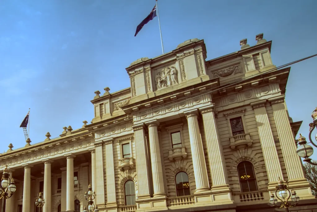 The sandstone colonnade and upper facade of Parliament House in Melbourne under a blue sky, with flags flying from the roof and ornate street lamps in front.