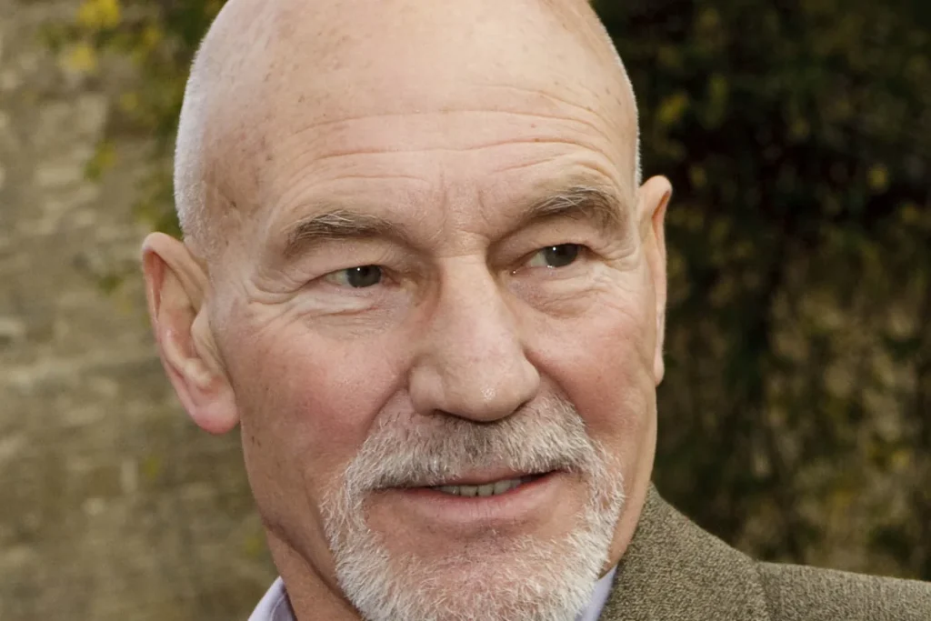Patrick Stewart, bald with a short white beard, looking slightly to the side in front of an ivy-covered stone wall.