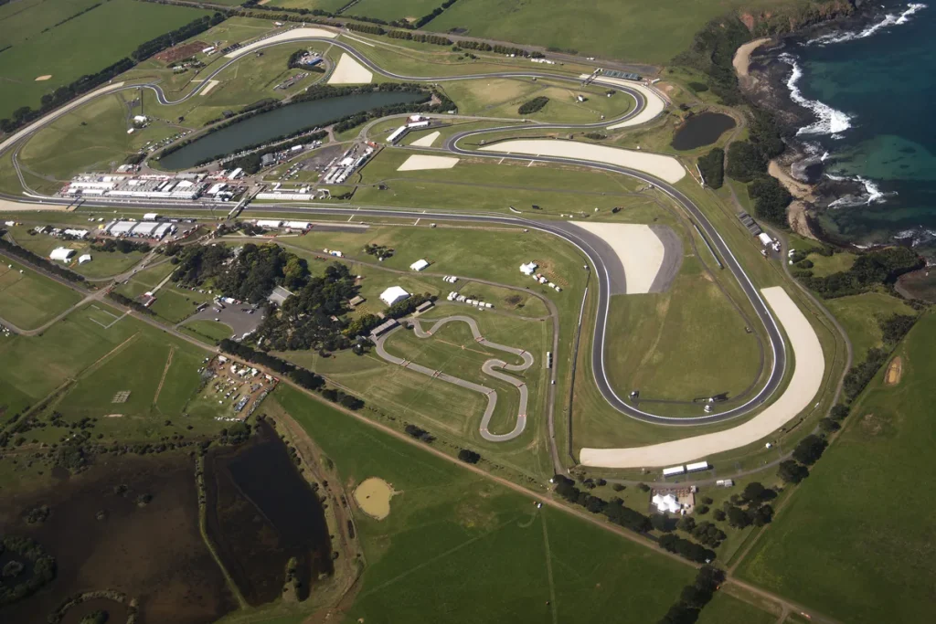 Aerial view of the Phillip Island race circuit: a grey ribbon of track with pale run-off areas winding through green paddocks beside a lake, the rocky coastline and blue sea at top right.
