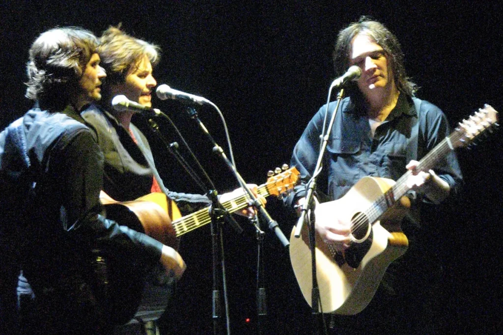 Three Powderfinger members on a dark stage, two sharing a microphone on the left and one on the right playing an acoustic guitar.