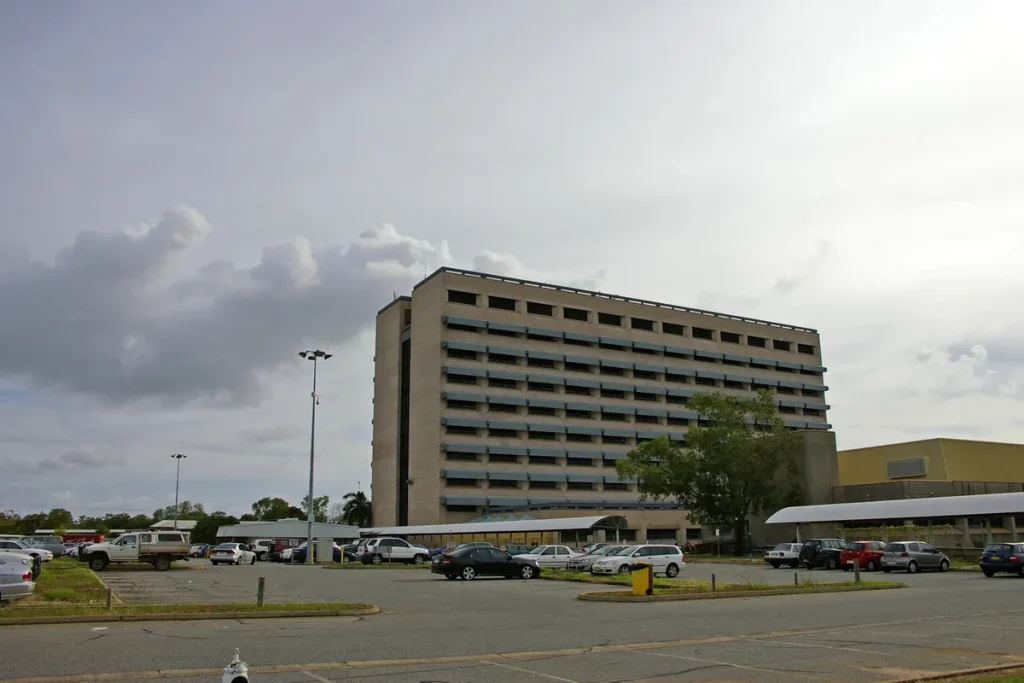The multi-storey main block of Royal Darwin Hospital under a cloudy sky, seen across an open car park with parked cars and a tree in front.