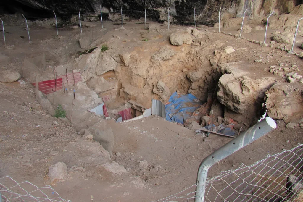 The floor of Shanidar Cave in Iraq, with an excavation trench cut into the earth and partly covered by blue tarpaulin and red sheeting, surrounded by a wire safety fence on metal posts.
