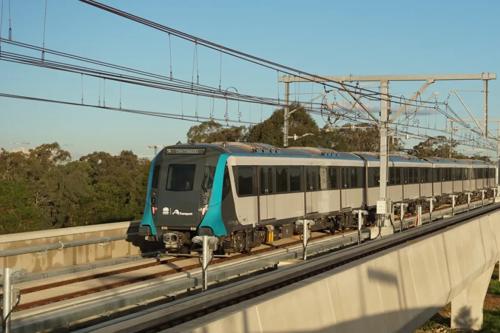 A grey and teal driverless Sydney Metro train on an elevated concrete viaduct under overhead wires, with gum trees and a clear evening sky behind.