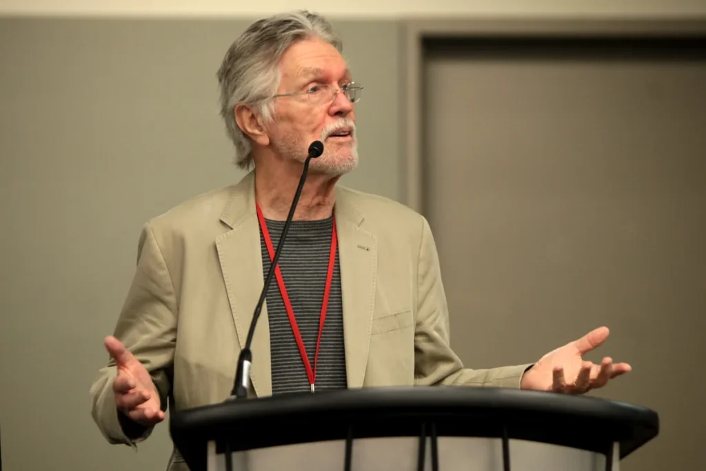 Tom Skerritt, with grey hair, glasses and a white beard, in a beige jacket over a striped top, speaking at a lectern with both hands open.