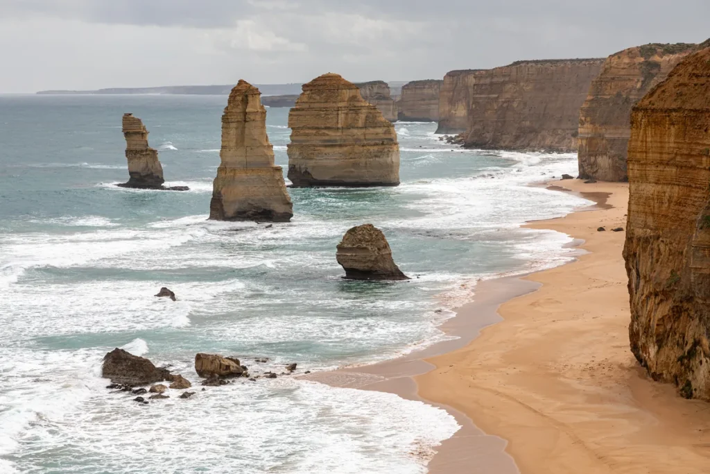 The Twelve Apostles limestone stacks rising from white surf off Port Campbell National Park, with a curving sandy beach and tall golden cliffs on the right under an overcast sky.