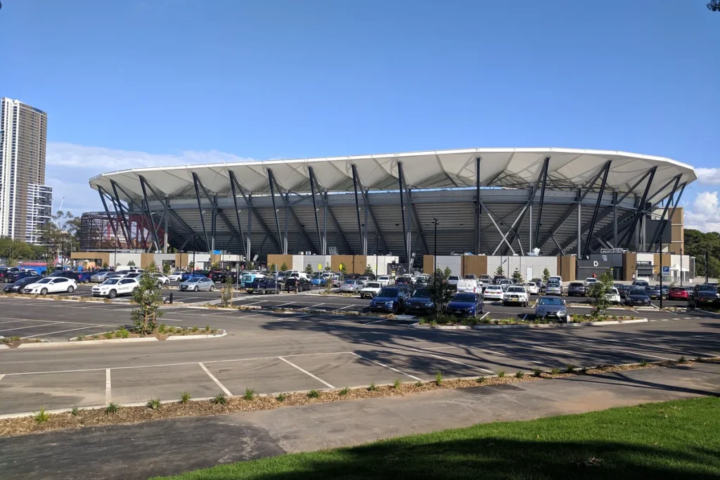 A modern stadium with a white wave-shaped roof on dark steel supports, seen across an open-air car park under a clear blue sky, with a residential tower at left.